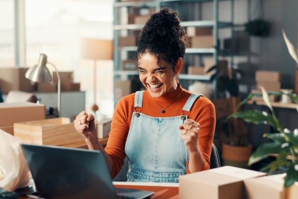 A happy woman raising her arms in delight while sat at her laptop.